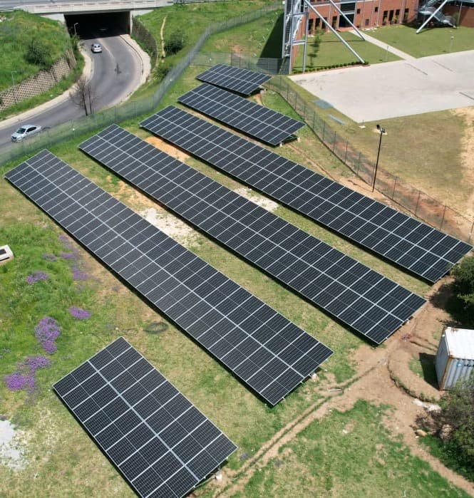 Aerial view of ground-mounted solar panel rows on a grassy slope beside a road